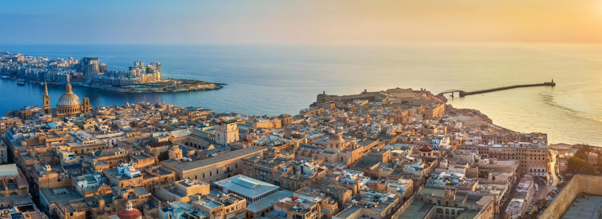 A panoramic aerial view of a coastal city at sunset with historic buildings, domed churches, dense urban architecture, a harbor with boats, and a long breakwater extending into the sea.