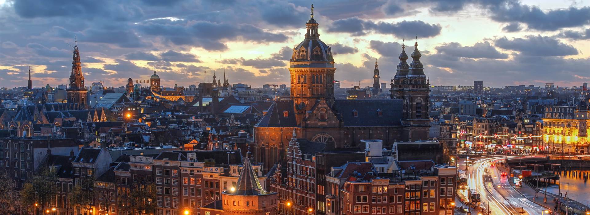 A panoramic view of Amsterdam at dusk, showcasing the historic architecture with illuminated street lights and buildings, a vivid sunset sky, and the bustling city life.